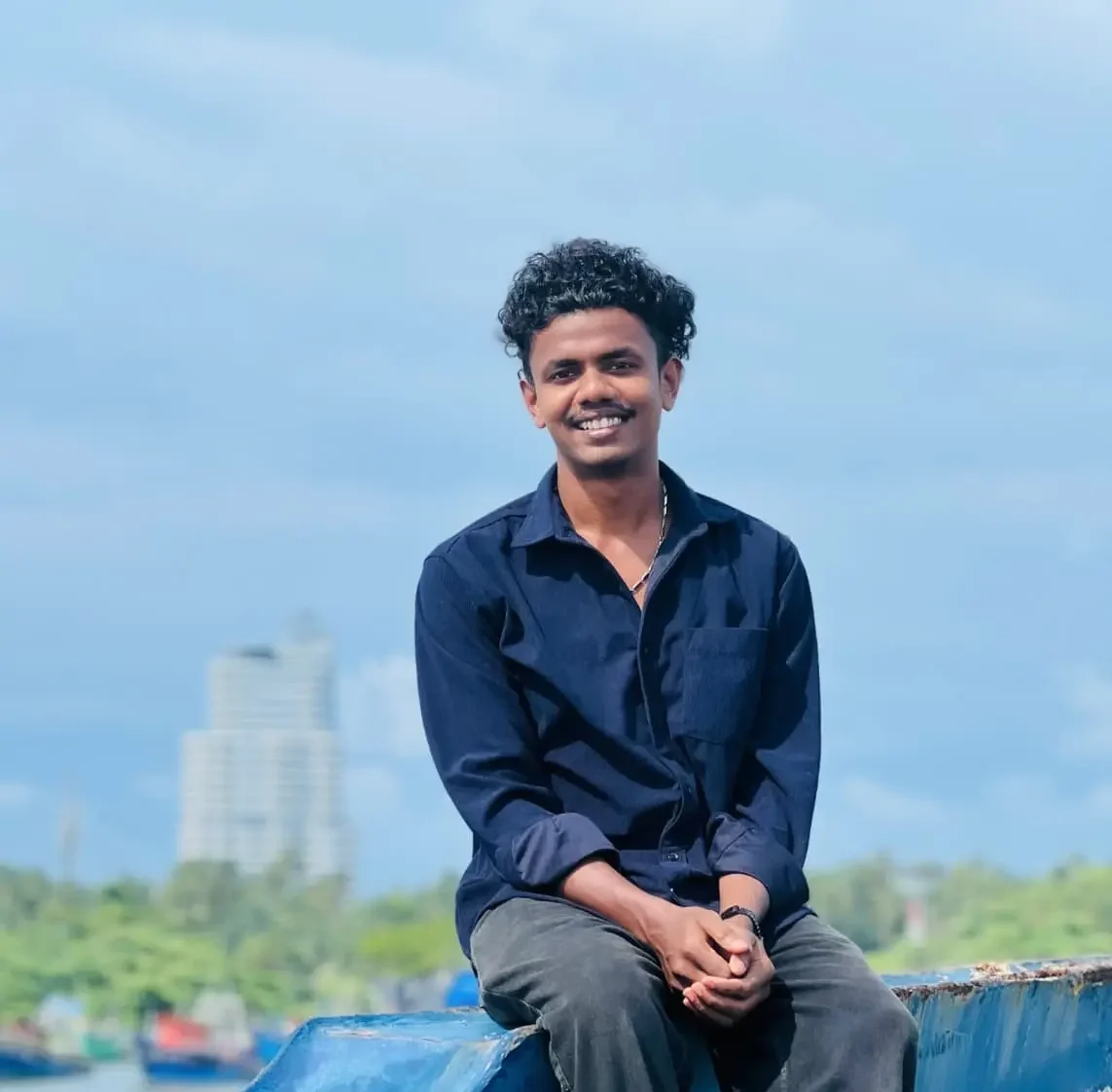 “A young man sitting on a blue boat edge, smiling confidently with a calm waterfront and cityscape in the background.”